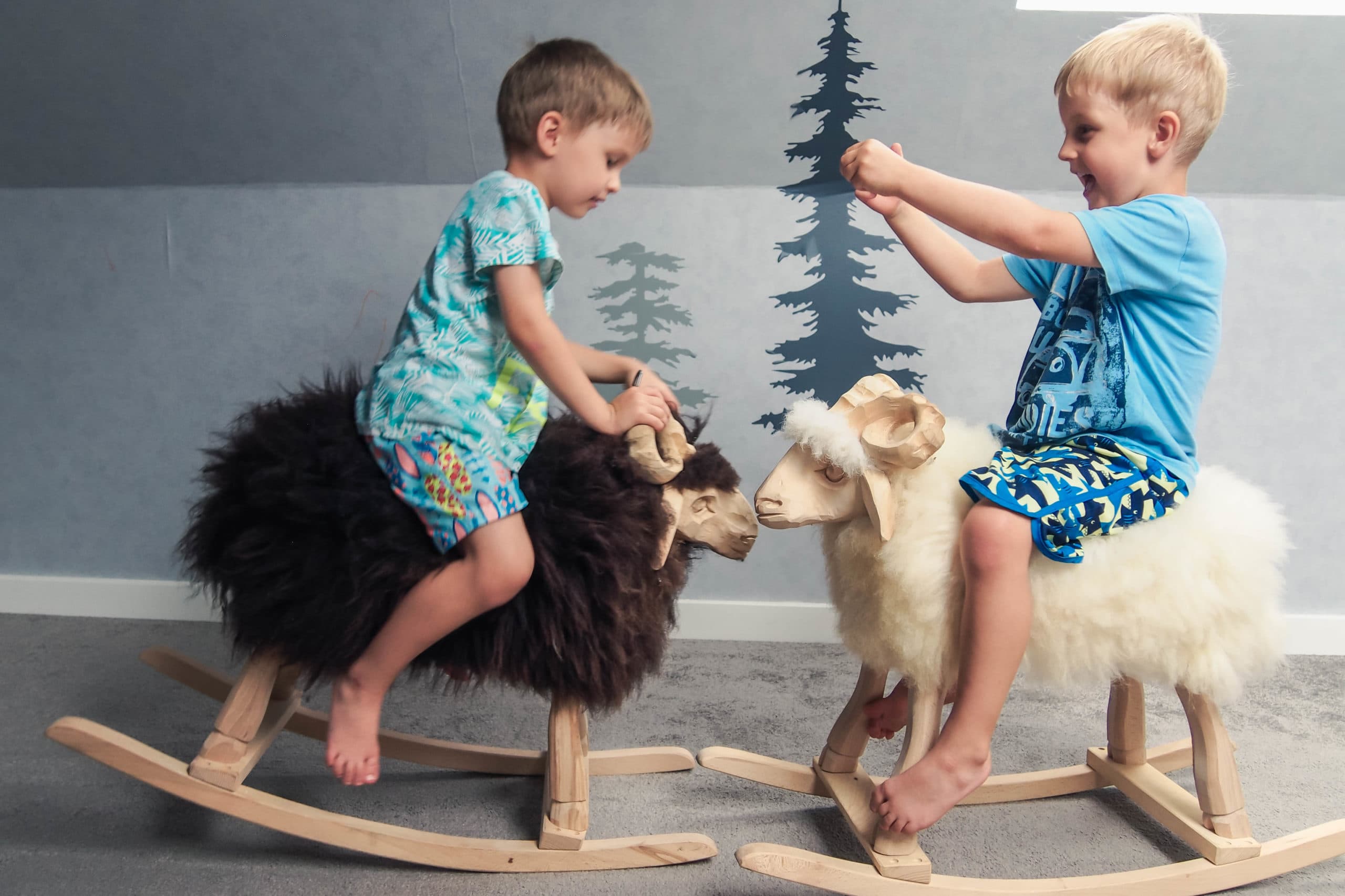 Two young boys play gleefully on furry rocking sheep indoors against wall mural