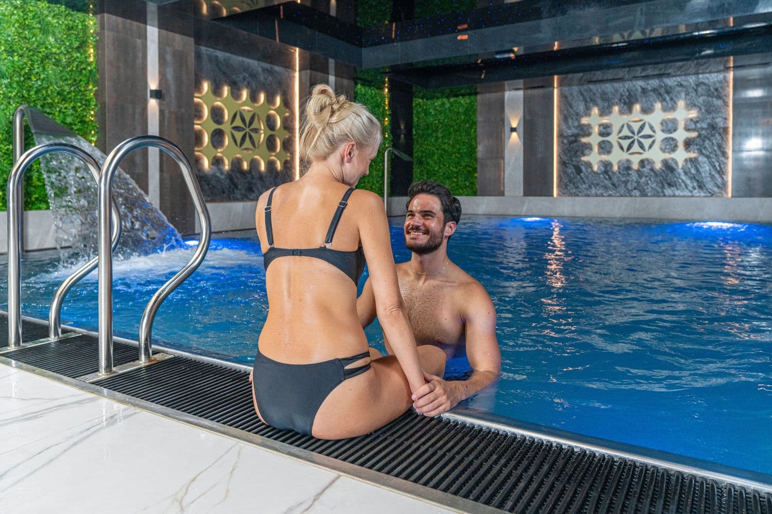 Woman sitting on pool edge and man smiling in water in indoor spa